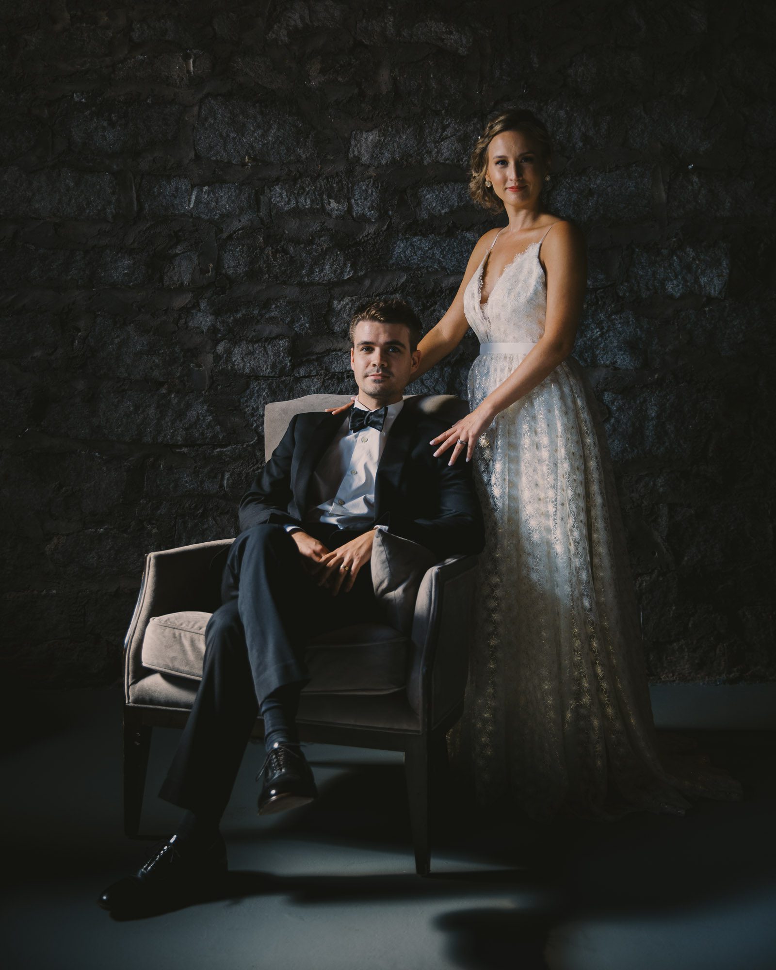 a bride wearing a white dress stands beside a groom wearing a black tuxedo who is seated in a chair in front of a stone wall on their wedding day at Atlanta's Terminus 330 wedding venue