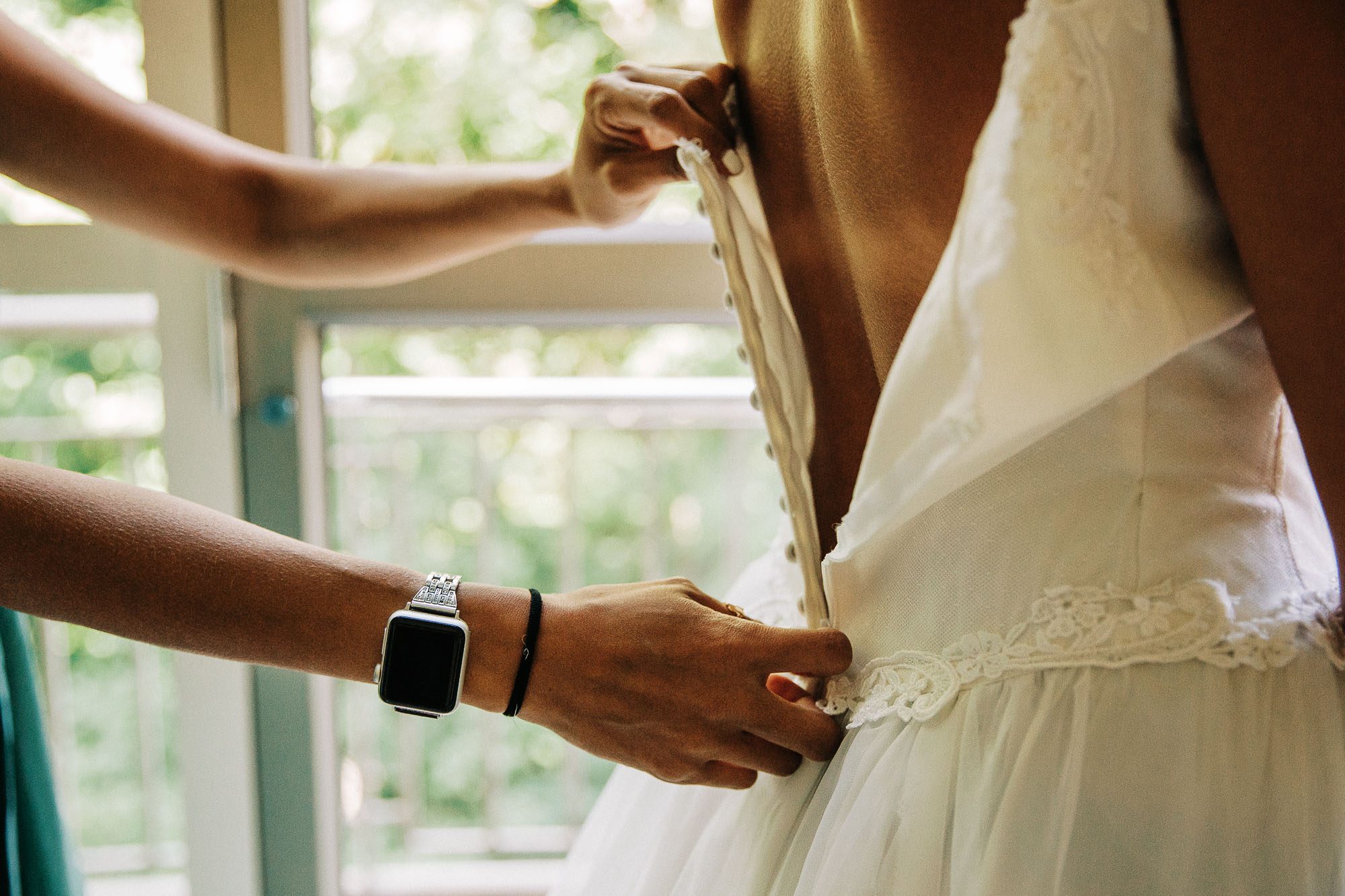 A close-up of the hand of the maid of honor buttoning the white wedding dress of the bride