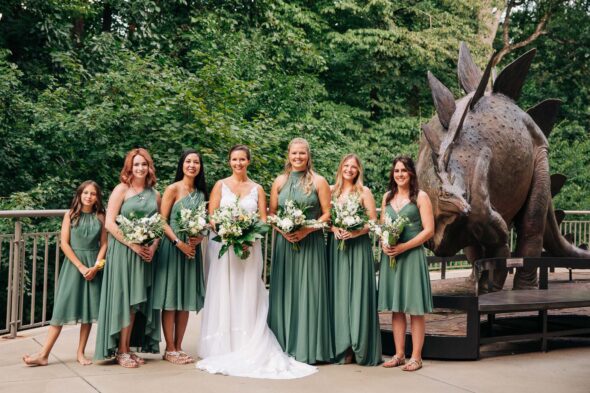 A bride and her bridesmaid stand holding their flowers next to a dinosaur sculpture at Fernbank science center