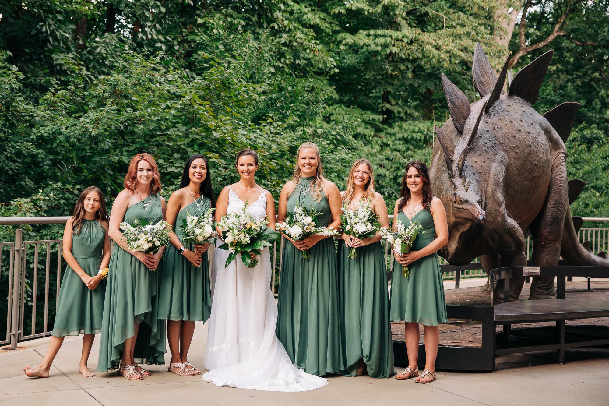 A bride and her bridesmaid stand holding their flowers next to a dinosaur sculpture at Fernbank museum