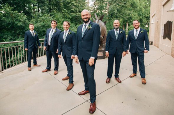 A groom and his groomsmen stand in front of a dinosaur sculpture at Fern Bank science cent