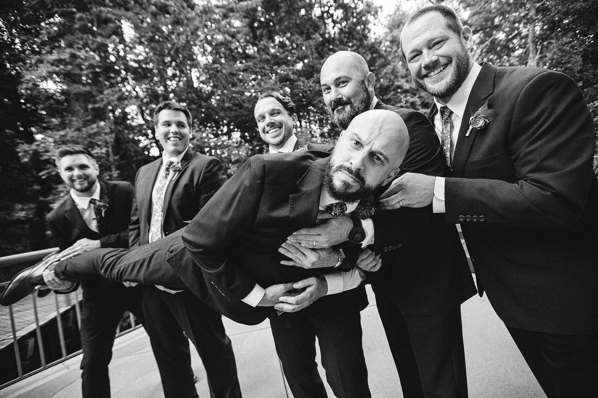 A black-and-white photo of a groom making a silly face while the groomsmen hold him in the air