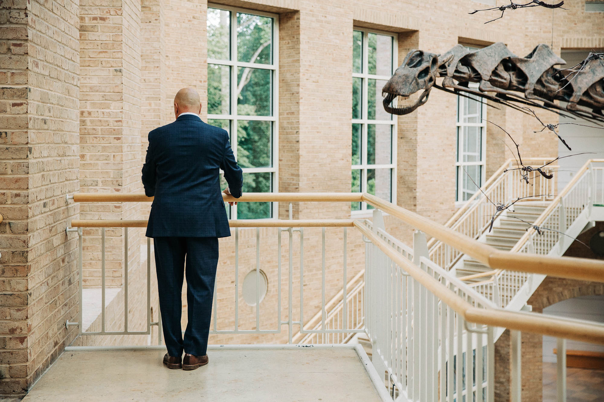 The father of the bride stands with his back to the camera waiting for his daughter on a balcony inside Fernbank museum 