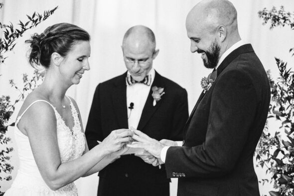 A close-up of a bride putting a ring on a grooms finger in black-and-white