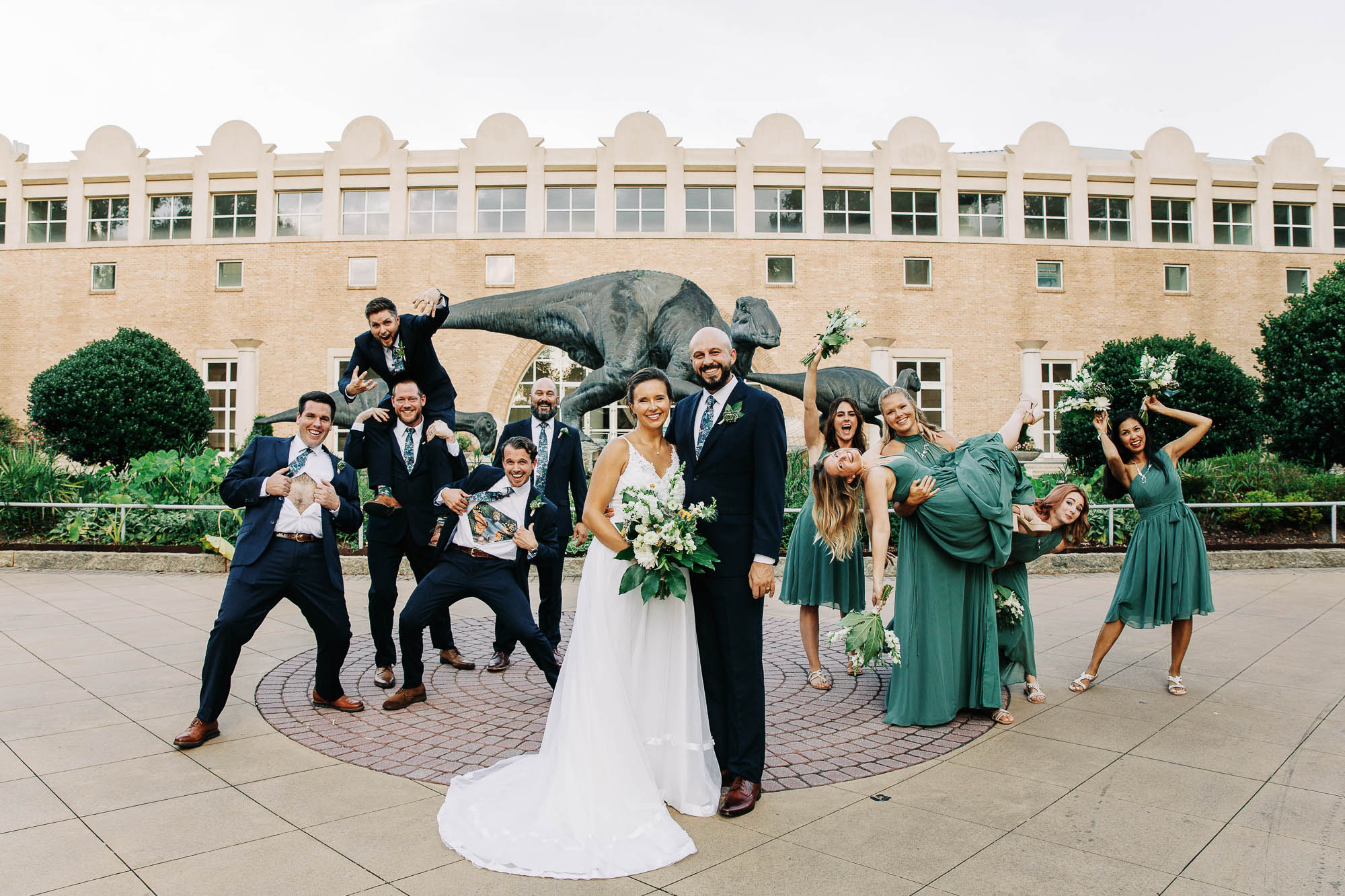 A silly photo of a wedding party and a bride and groom standing in front of Fern Bank museum with the dinosaur sculpture behind them