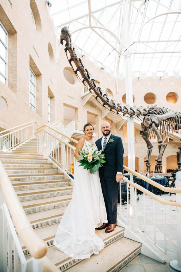 A bride and groom pose on the stairs underneath the giant dinosaur skeleton in the atrium at Fern Bank science center