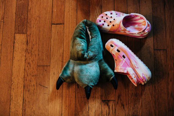 One dinosaur foot bedroom slipper and two pink croc shoes lie on a hardwood floor