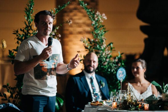 A best man wearing a funny T-shirt gives a speech while the bride and groom look on