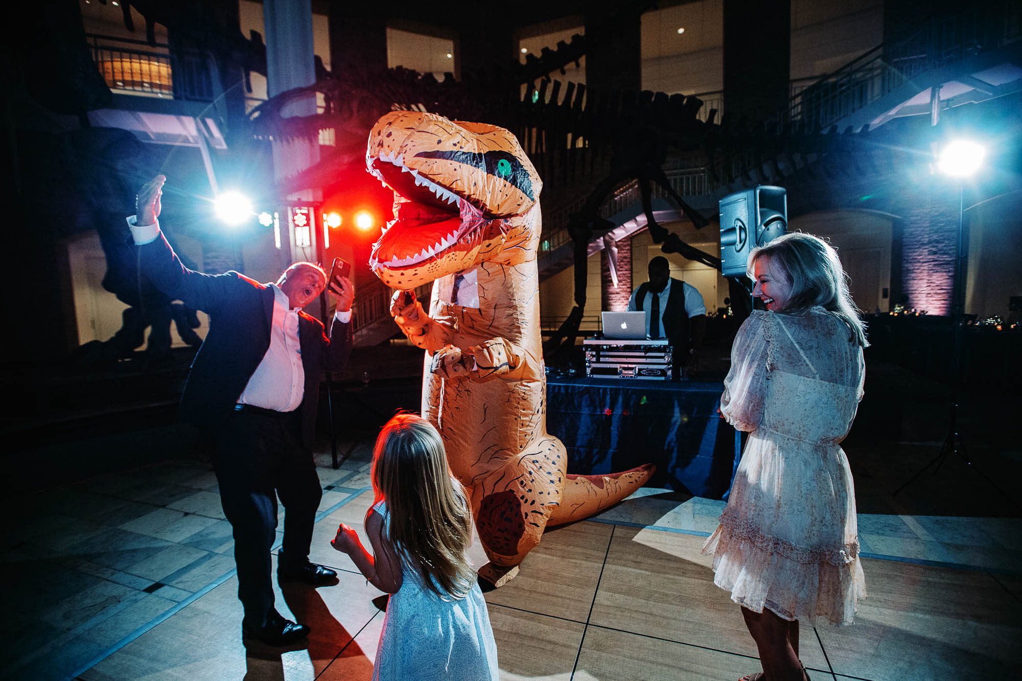 A colorfully lit photo of a person in a dinosaur costume on the dance floor with other guests at a wedding at Fern Bank museum
