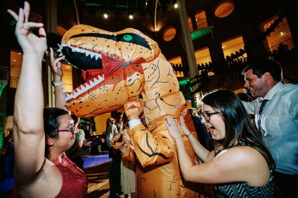 A person in a dinosaur costume and two women dance on the dance floor at a wedding