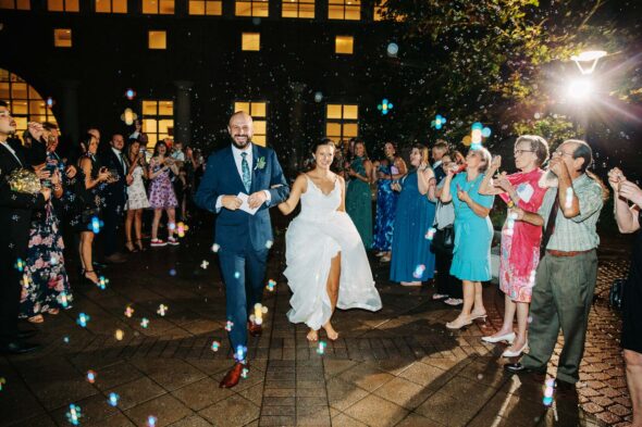 A bride and groom walk out of their wedding reception and ask their guest lineup on either side of them and blow bubbles in the rain