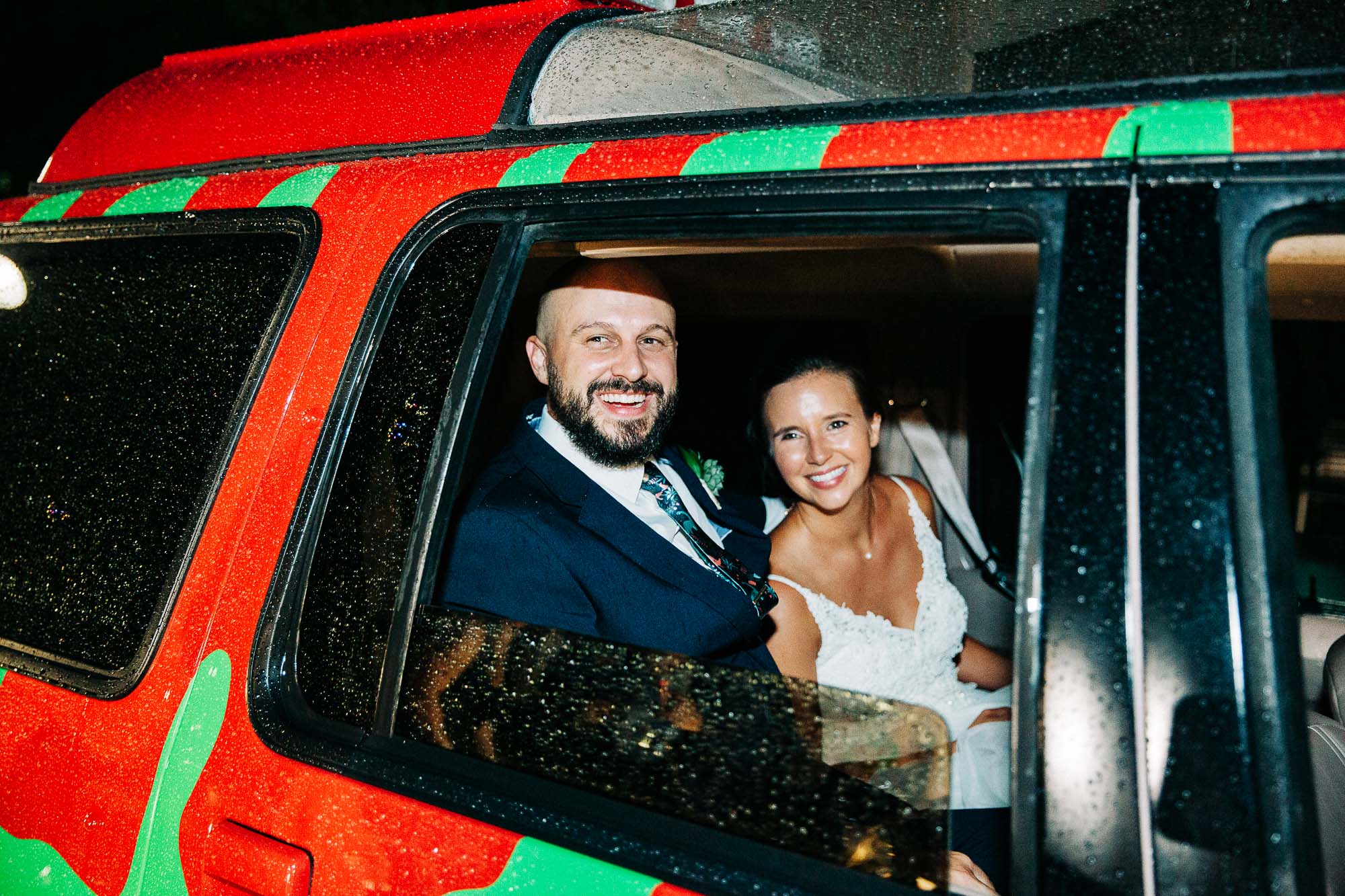 Close up photo of a bride and groom looking out of the window of their getaway car and smiling