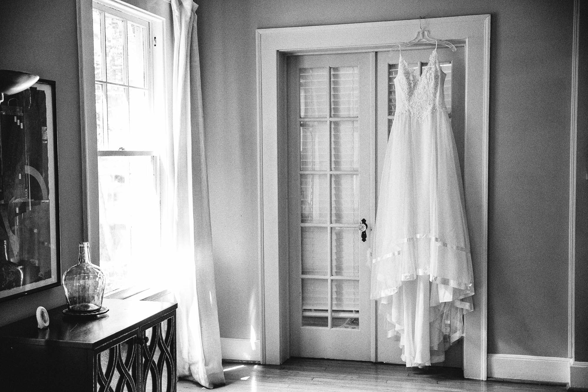 Black and white photo of a white wedding dress hanging on a door next to a window