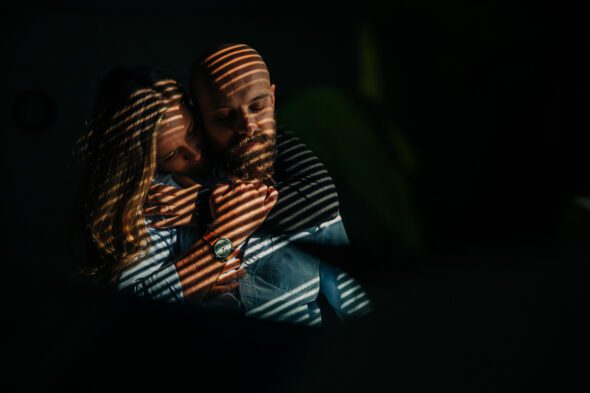 a man and woman embrace in light cast by a window in their home for their lifestyle engagement photo