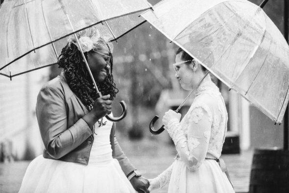 black and white photo of two brides in wedding dresses holding umbrellas in the rain seeing each other for the first time on their wedding day