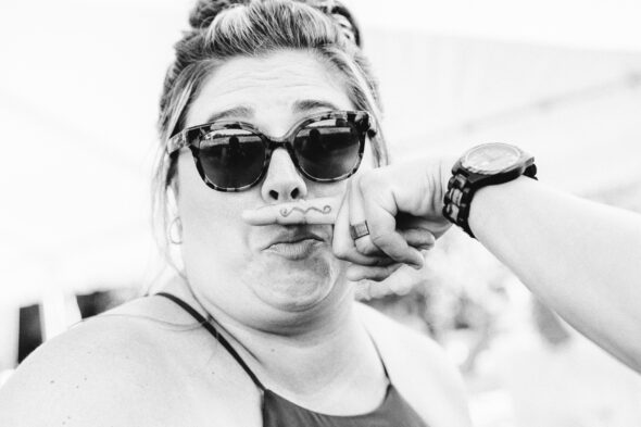 funny photo of a woman wearing sunglasses making a silly face while a person off camera holds their finger with a mustache tattoo in front of her lip