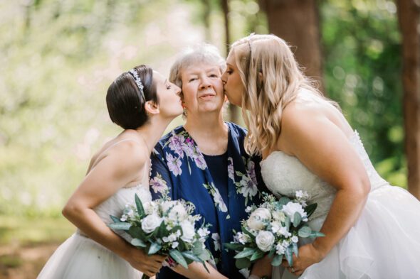 two brides wearing white dresses kiss their grandmother