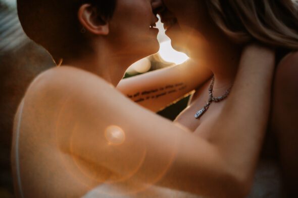 very close photo of two brides wearing white wedding dresses kissing at sunset with camera lens flare