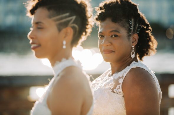 two black brides wearing white wedding dresses stand together with one looking at the camera and one looking to the left