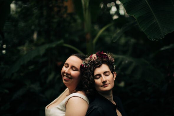 a couple poses in the tropical room at the Atlanta botanical garden. it's their wedding day and one is wearing a white dress and the other is wearing a flower crown and black clothing