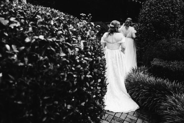 two brides wearing white wedding dresses walk away from the camera between giant shrubs at Primrose Cottage in Roswell Georgia