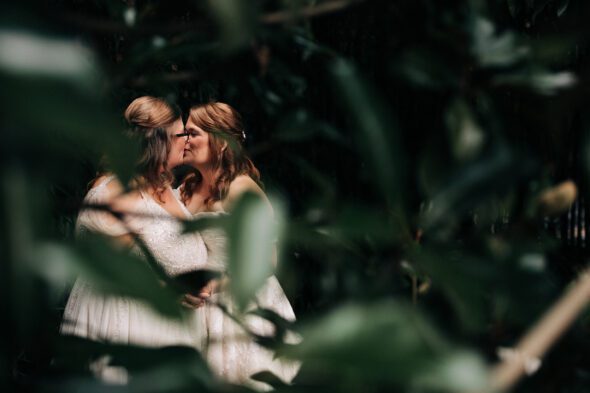 two brides wearing white wedding dresses kiss behind a tree at Primrose Cottage in Roswell Georgia