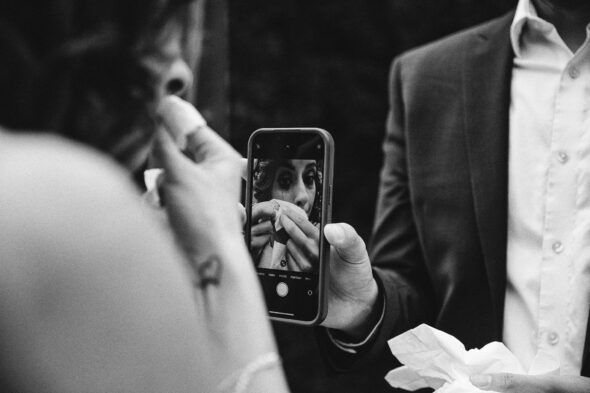 black and white photo of a bride looking into a cellphone camera to wipe her tears