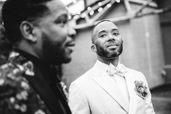 black and white photo of a black groom in a white tuxedo looking at his husband