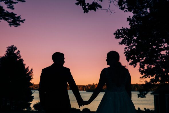 a couple in silhouette holds hands at sunset in front of a lake with a colorful sky