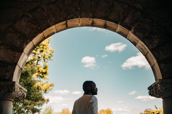 a black groom wearing a white tuxedo stands with his back to the camera and looks to the right on the porch of Rhodes Hall in Atlanta