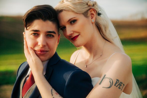 a transmasc nonbinary person wearing a blue suit and their bride wearing a white dress embrace and look at the camera on their wedding day