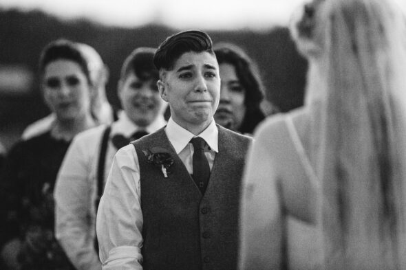 a transmasc nonbinary person in a vest and tie looks emotionally at their bride during their sunset wedding ceremony