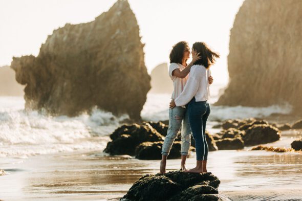a lesbian couple embraces while standing on a rock at the beach for their engagement photos