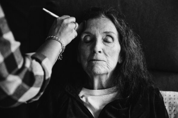 black and white photo of an older woman with her eyes closed as a hand applies makeup to her face