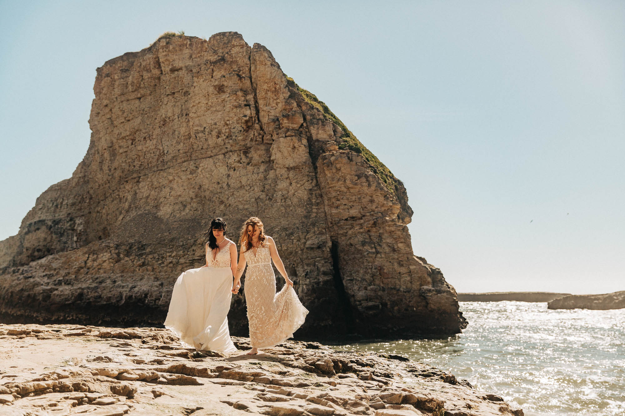 two brides walk on the rocks in white wedding dresses at shark fin beach