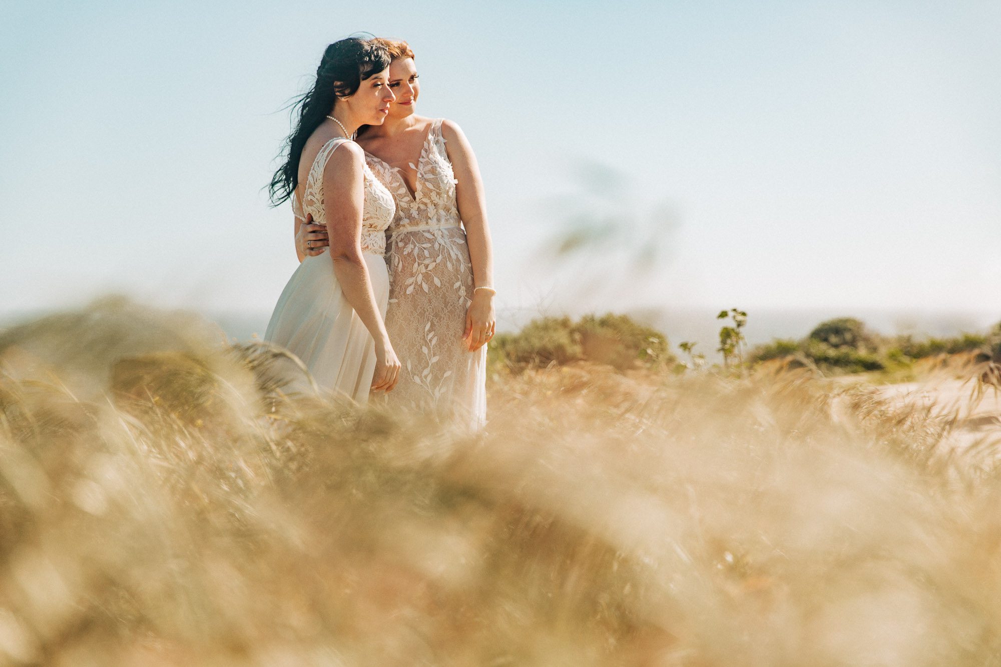 two brides in white wedding dresses stand in the grass above davenport beach