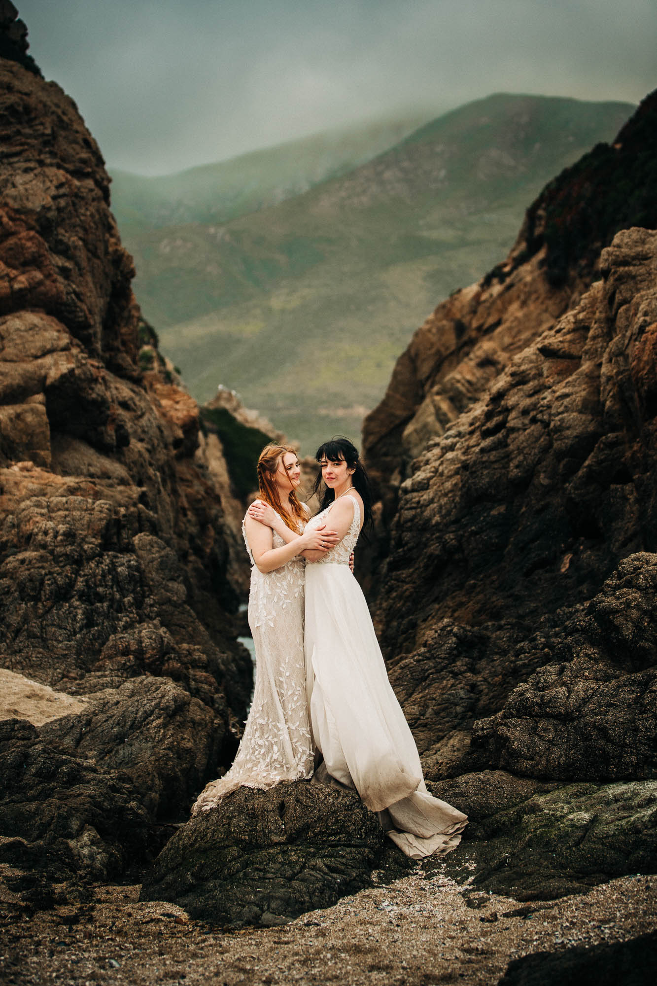 two brides in white wedding dresses stand on the rocks at garrapata state park
