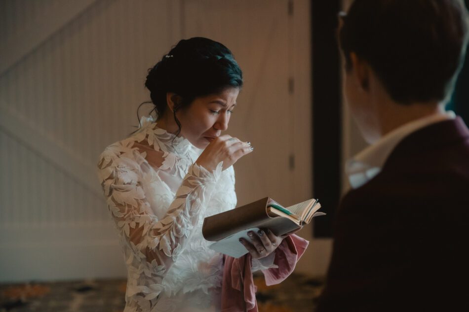 a bride wearing a white wedding dress cries while she opens the gift her partner has given her during their first look at their LGBTQ Virginia wedding
