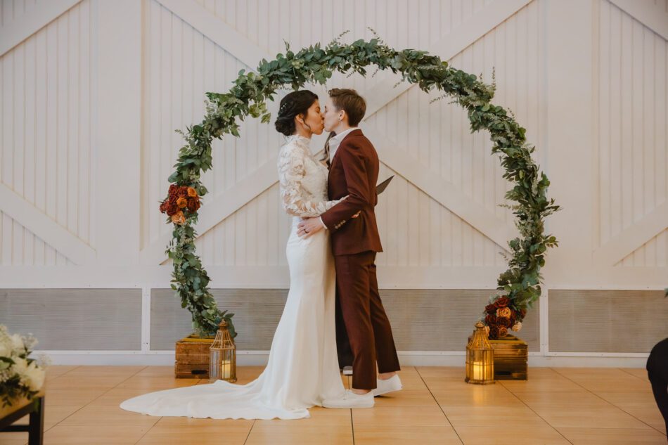 a woman in a burgundy Bindle and Keep suit kisses her wife wearing a Maggie Sottero white wedding dress at the end of their wedding ceremony in Virginia