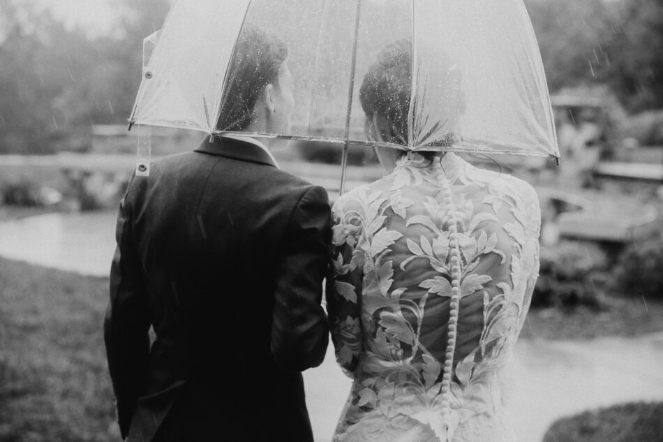 a woman in a suit and a woman in a white wedding dress under a clear umbrella walk away from the viewer on their rainy wedding day at their LGBTQ Virginia wedding