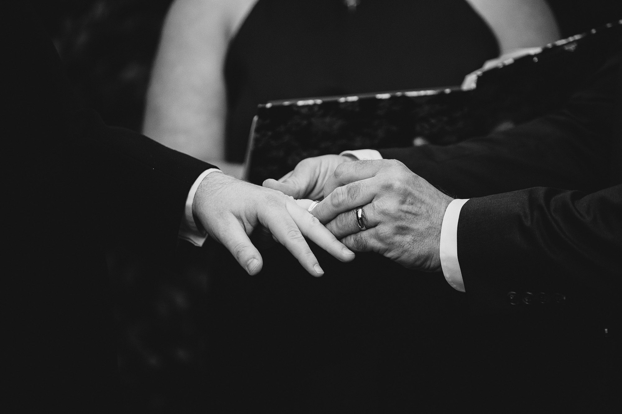 moody black and white photo of just the hands of two grooms as they exchange wedding rings at their rainy backyard Atlanta wedding ceremony