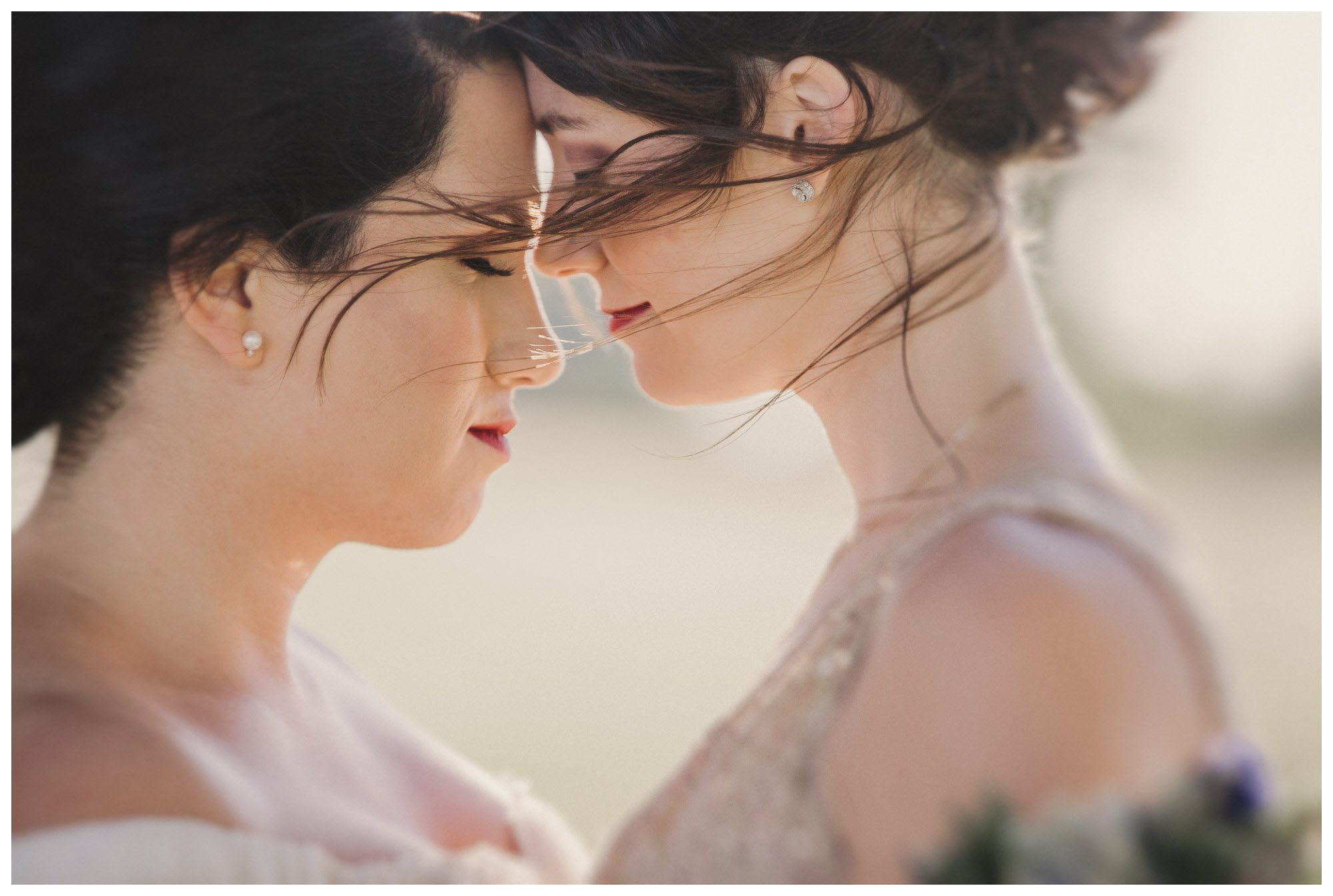 two brides lean close as their hair blows in the wind on their wedding day