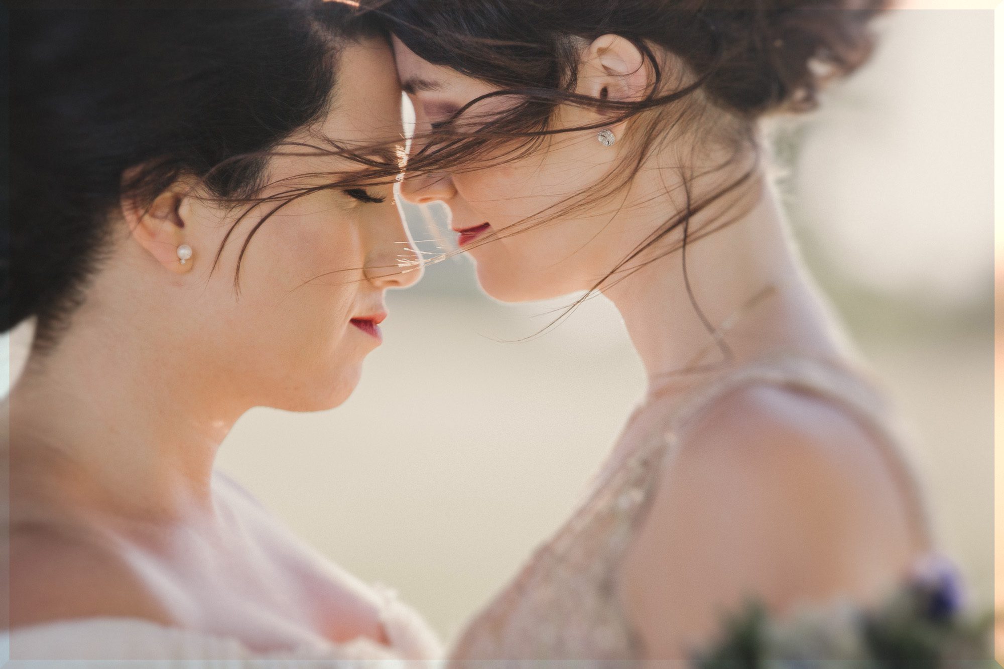 lesbian couple in white wedding dresses lean their foreheads against each other as the wind blows their hair gently on their wedding day