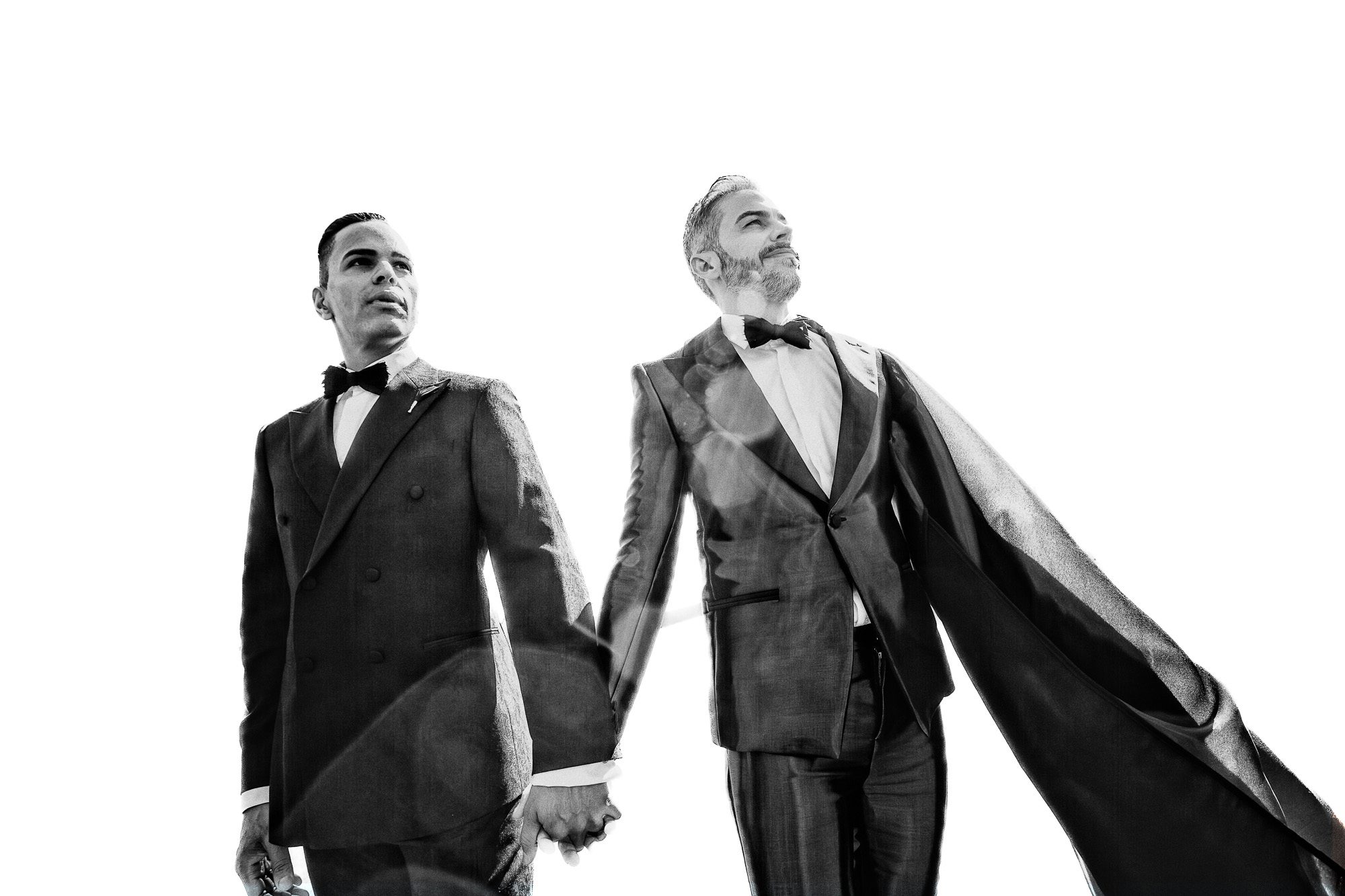 black and white photo of two men wearing black suits hold hands and look up and away from the camera for their wedding portrait