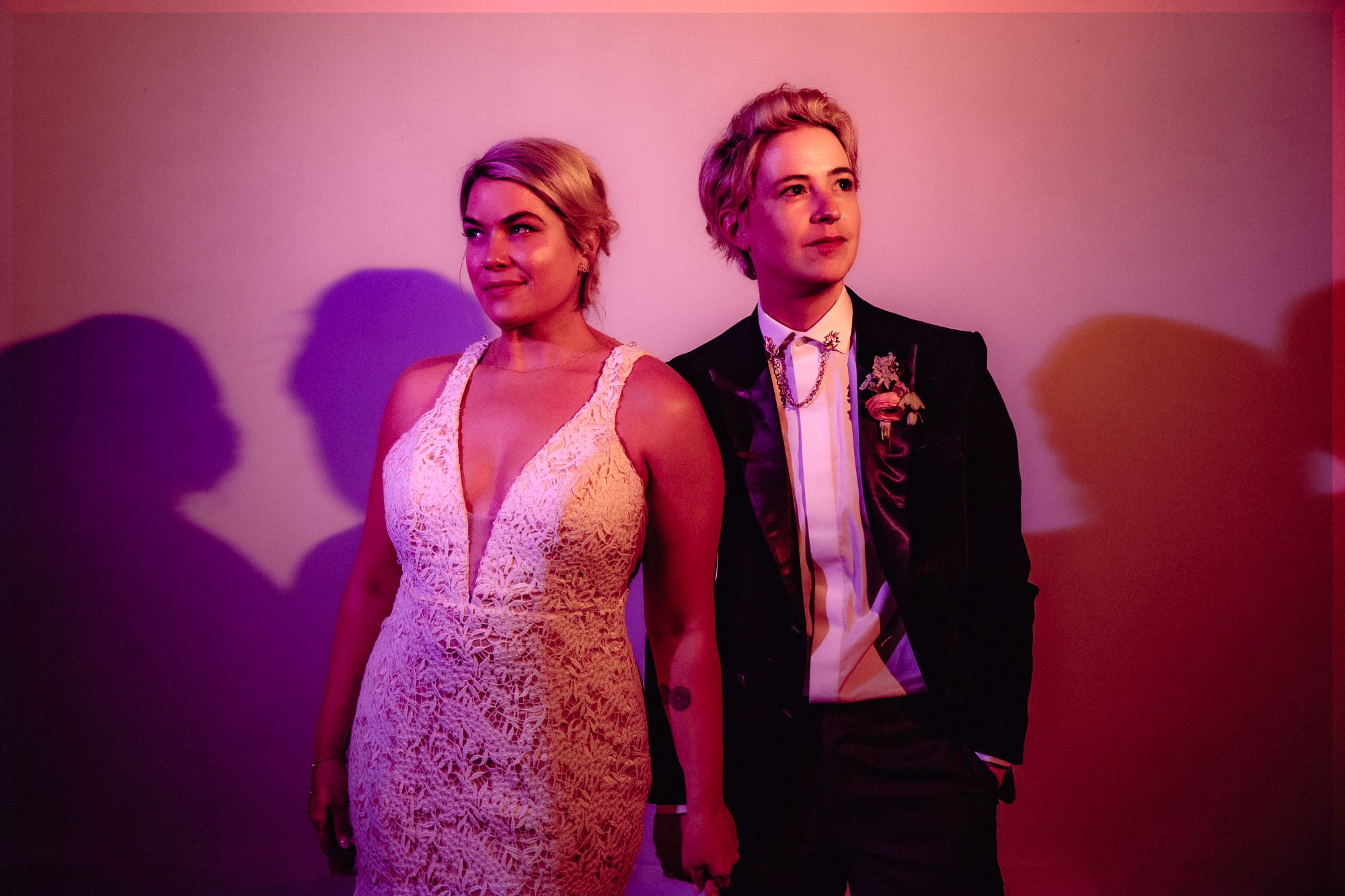 colorfully pink photo of an LGBTQ couple wearing a white wedding dress and a dark suit holding hands and looking up and away from the camera at their Atlanta wedding portrait at Westside Warehouse