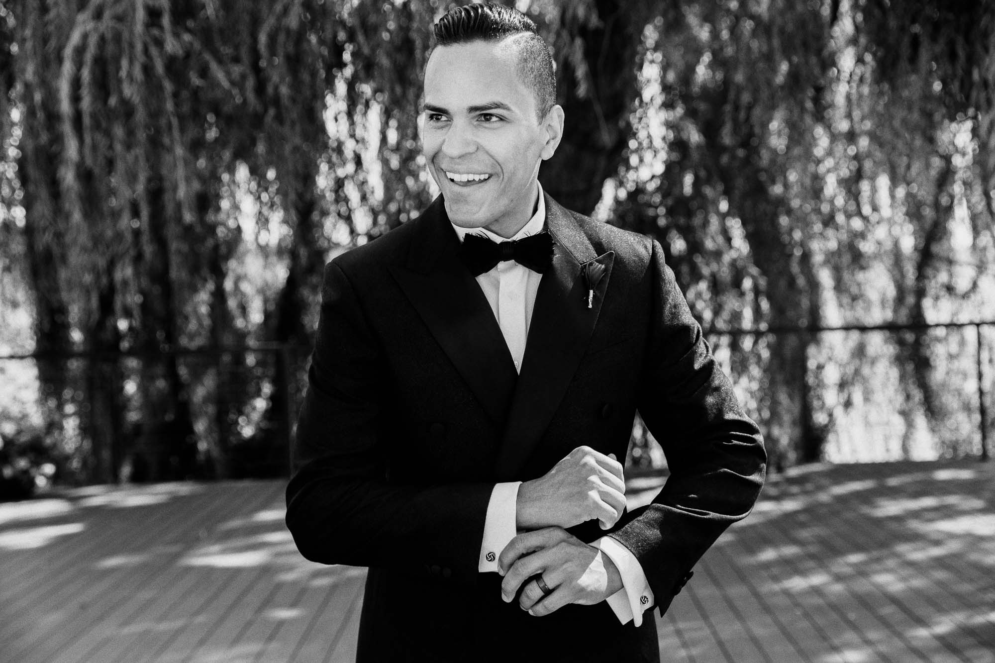 black and white photo of a groom in a black suit laughing and straightening his cufflinks
