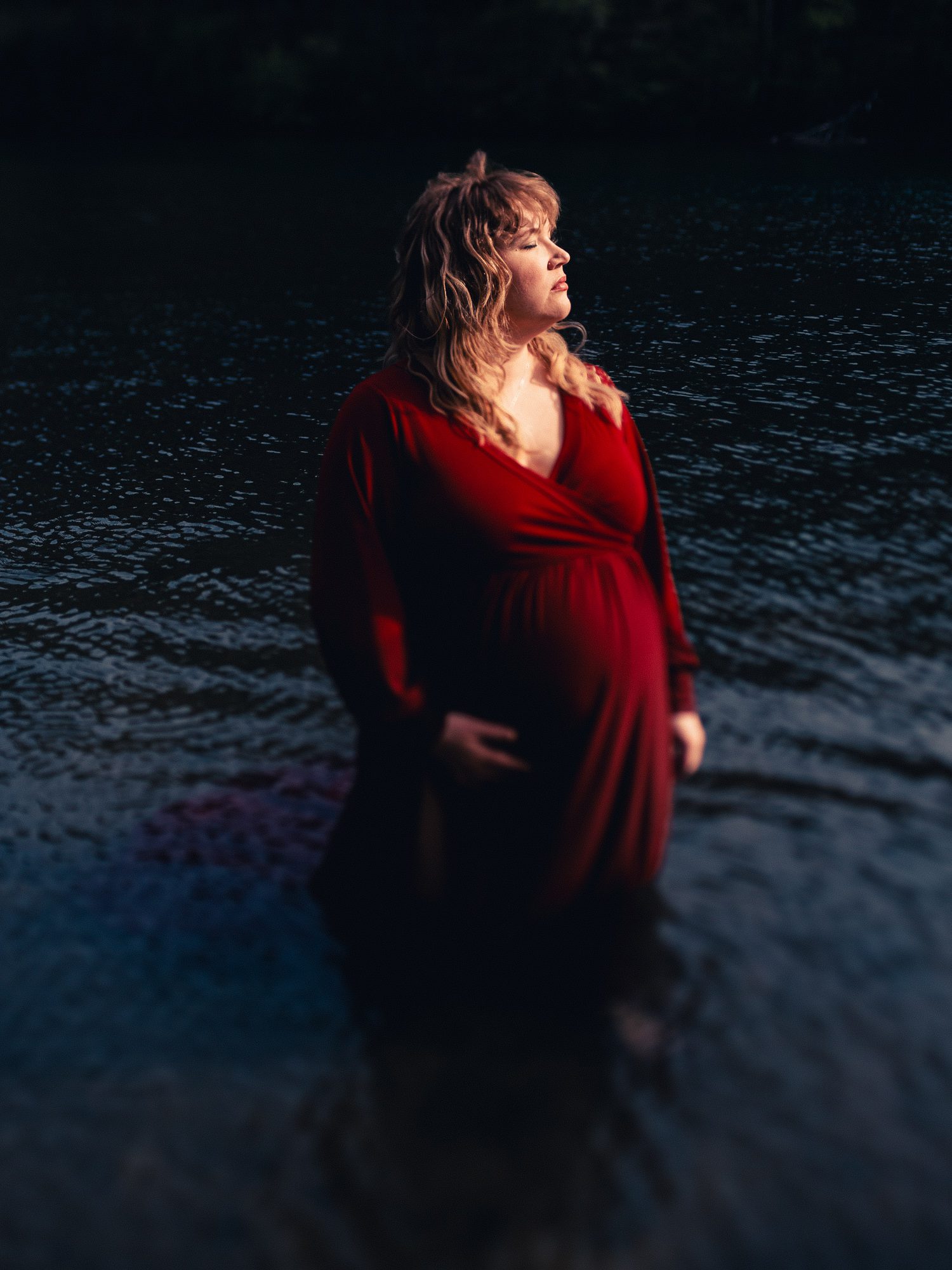 a pregnant woman wearing a flowing red dress stands knee deep in the water at Lake Rabun facing the warm setting sun