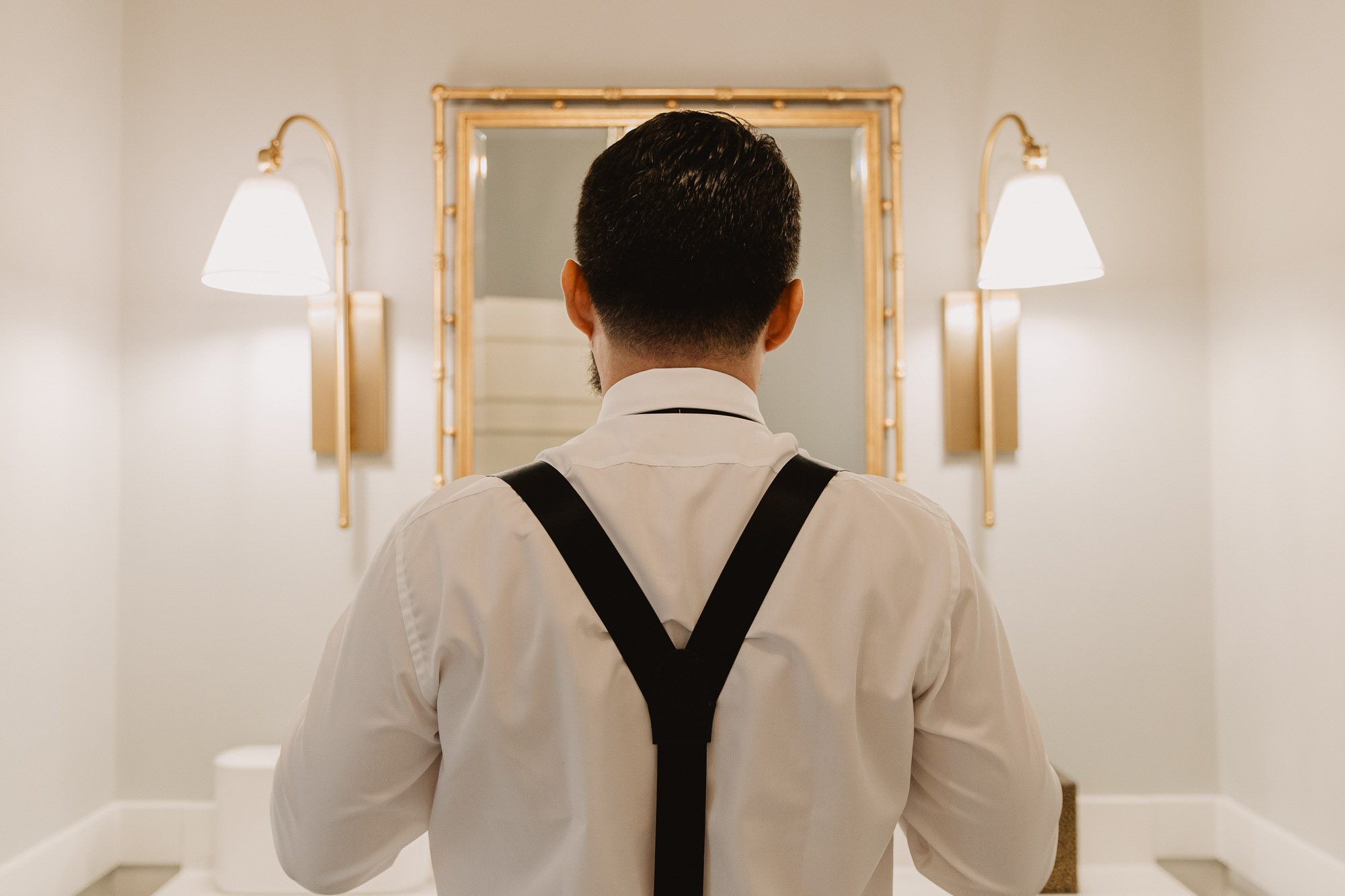 A groom wearing suspenders stands facing a mirror in the bathroom at Atlanta's historic Wimbish house on peach tree