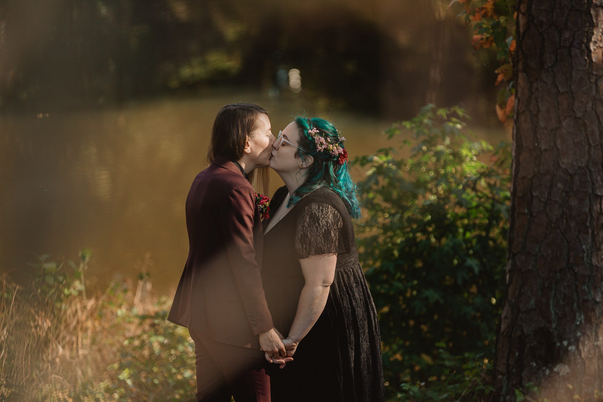 a bride with beautiful blue hair wearing a black wedding dress embraces her nonbinary partner wearing a deep purple suit while standing in the sunlight in a wooded area beside a lake on their wedding day