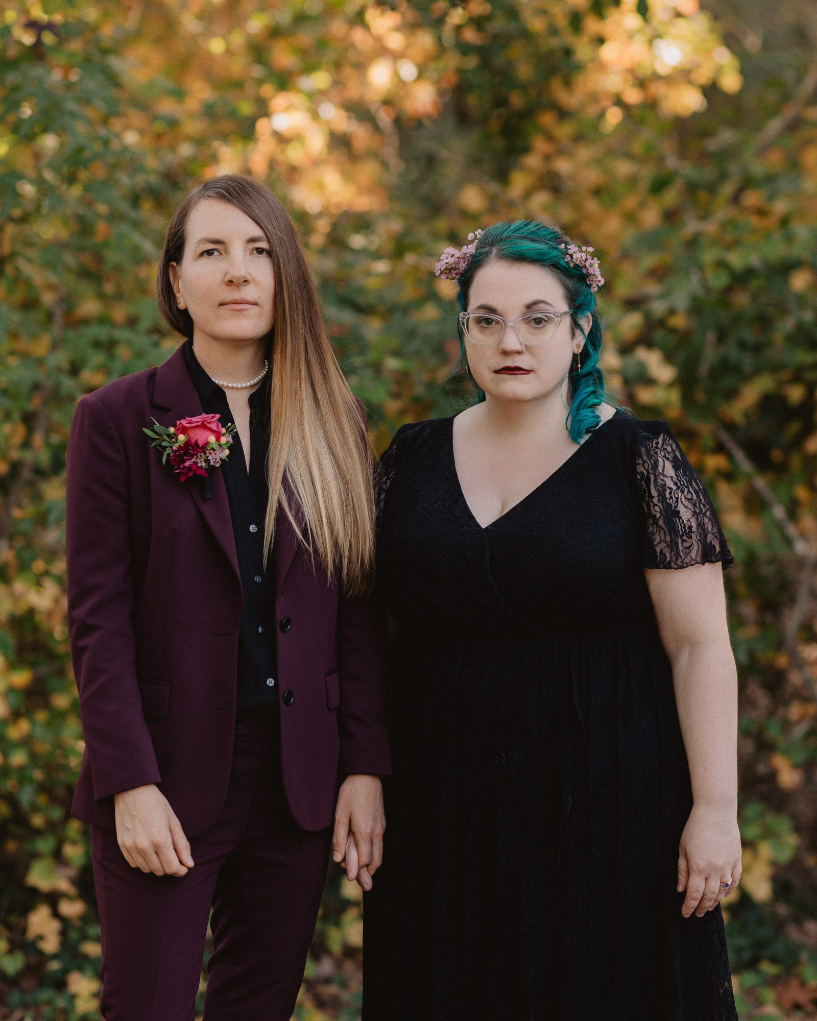 wedding portrait of a gorgeous queer couple on their fall wedding day in Atlanta, on the left is a nonbinary marrier wearing a deep purple suit with a bright red boutonniere and on the right is a bride with teal hair wearing a black wedding dress